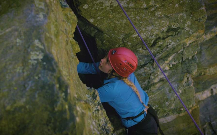 A young person wearing safety gear is secured by ropes as they navigate a crack in a rock wall they are climbing. 